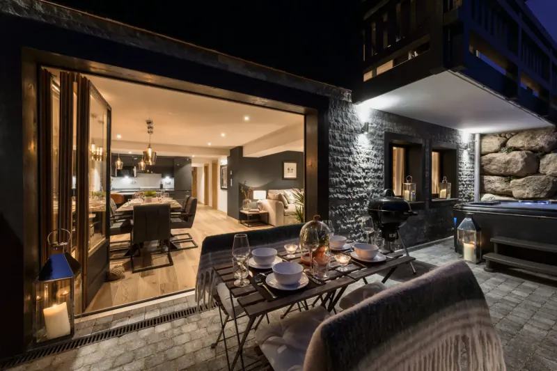 Evening view of Hunter Chalets terrace with set dinner table, open doors to modern kitchen and living room, hot tub, mountainside.