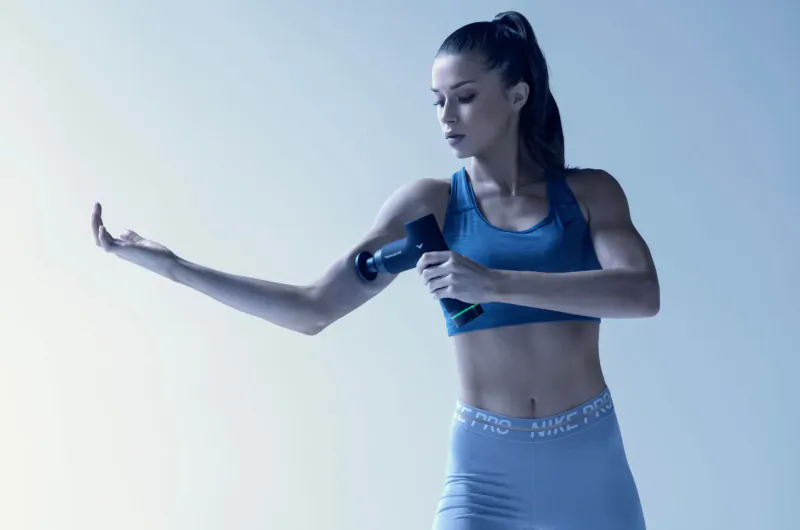Athletic woman in blue workout gear using black Hypervolt GO massage gun on her arm against blue background