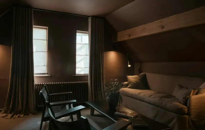 Cozy attic lounge with beige sofa, wooden chairs, radiator, plants, and draped windows in warm lighting