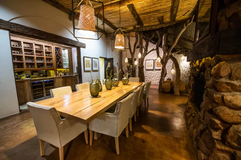 Elegant wooden dining room with long table, white chairs, wine cabinets, tree branch decor, and stone fireplace at Tswalu Tarkuni.