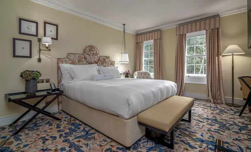 Elegant hotel bedroom with floral-upholstered king bed, bedside lamps, flowers, and window views.