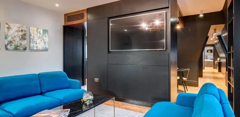 Modern hotel lounge with blue velvet sofas, glass coffee table with magazines, large TV on dark wall, and abstract art.