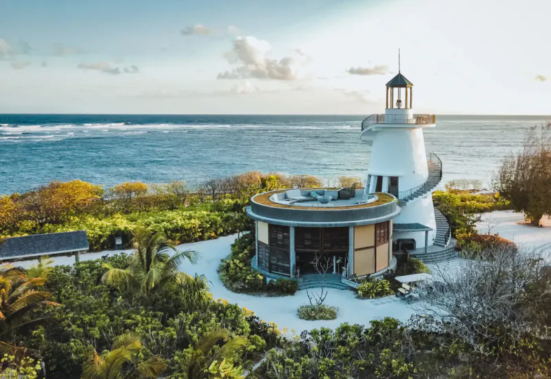 Modern white spiral lighthouse and pavilion on tropical island surrounded by lush greenery, beach, and ocean at sunset.
