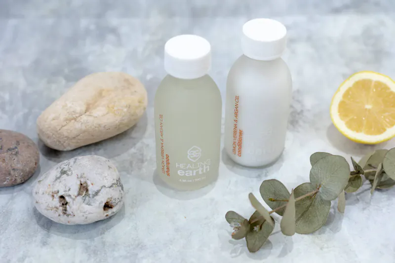 Two white glass bottles labeled 'Healing Earth' with clear and milky liquids, beside rocks, lemon slice, and eucalyptus on gray surface.