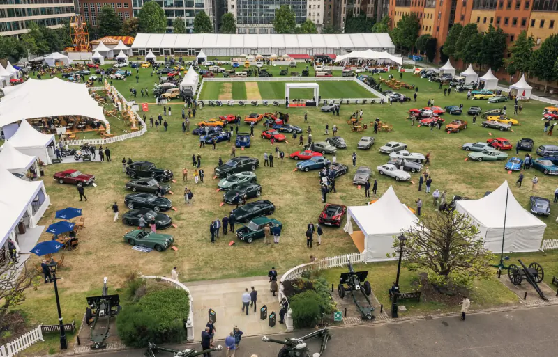 Aerial view of London Concours 2024 car show with classic V12 vehicles on grass amid white tents and crowds.