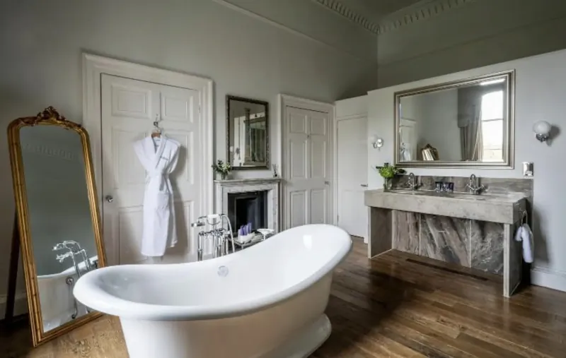 Elegant bathroom in Bailbrook House Hotel with white freestanding bathtub, gold mirror, white robe on door, and stone vanity.