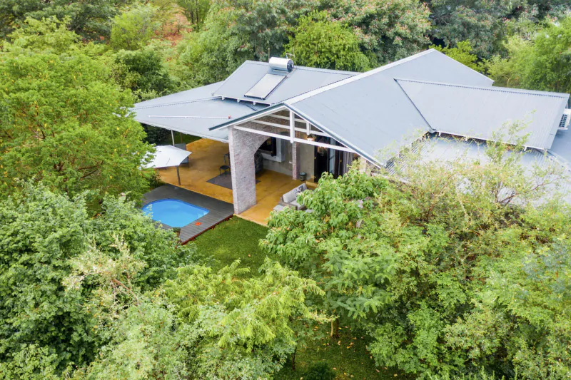 Modern house with blue roofs, yellow deck, small pool, and umbrella, surrounded by lush green trees near Victoria Falls.