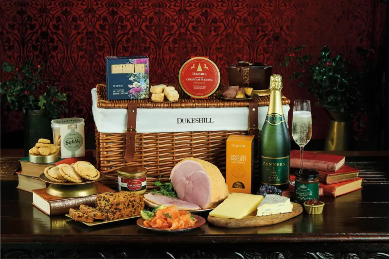 Dukeshill Christmas hamper basket with champagne bottle, ham, cheese, biscuits, chocolates on wooden table against red damask backdrop.