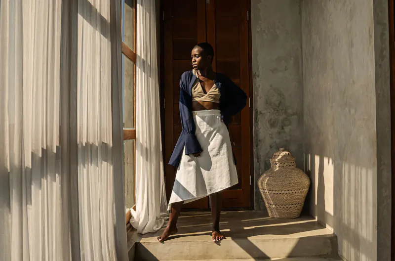 Black woman in beige bralette, white skirt, navy open jacket stands confidently by sheer-curtained door with woven pot, resort wear