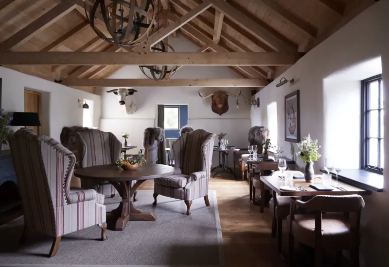 Cozy luxury lounge in rustic beamed room with striped armchairs, round table of fruit, antlers, and large windows.
