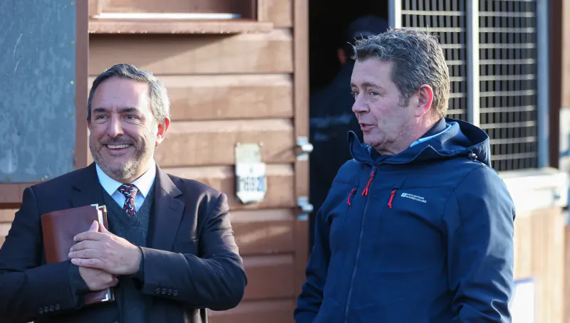 Two men, one in suit holding book and one in blue jacket, stand smiling outside wooden stable at Tattersalls horse ring.