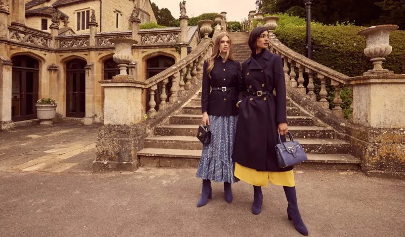 Two women in navy trench coats, skirts, boots and handbags stand on steps before grand stone manor house.