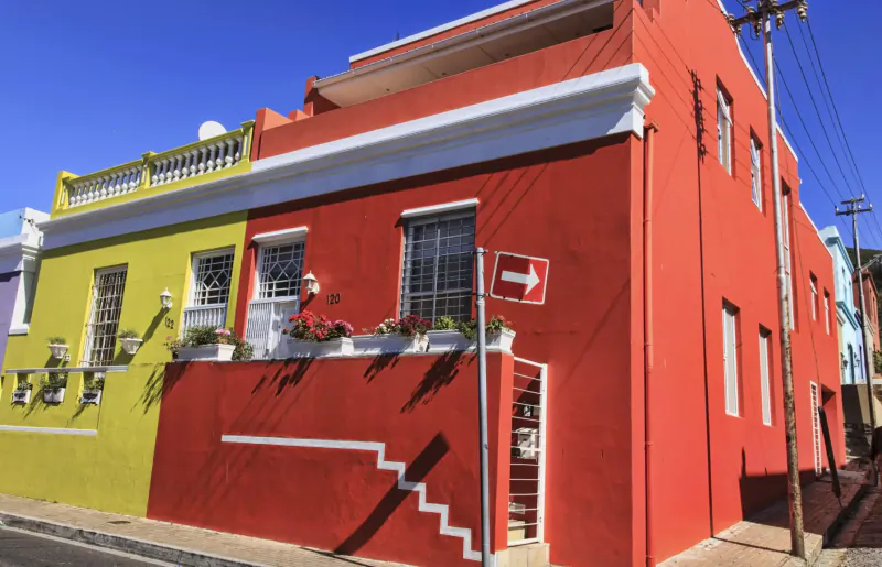 Colorful yellow and red Bo-Kaap houses with flower boxes, balcony railings, and red arrow sign under blue sky.
