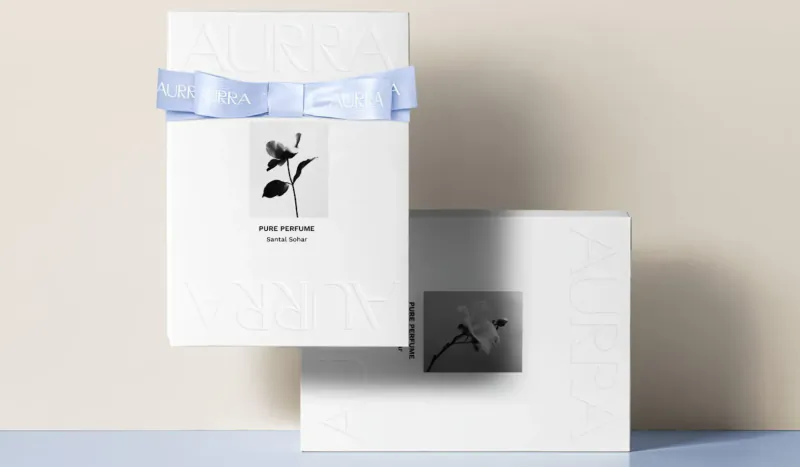 White AURRA PURE perfume box with blue ribbon bow and black leaf image, angled on surface