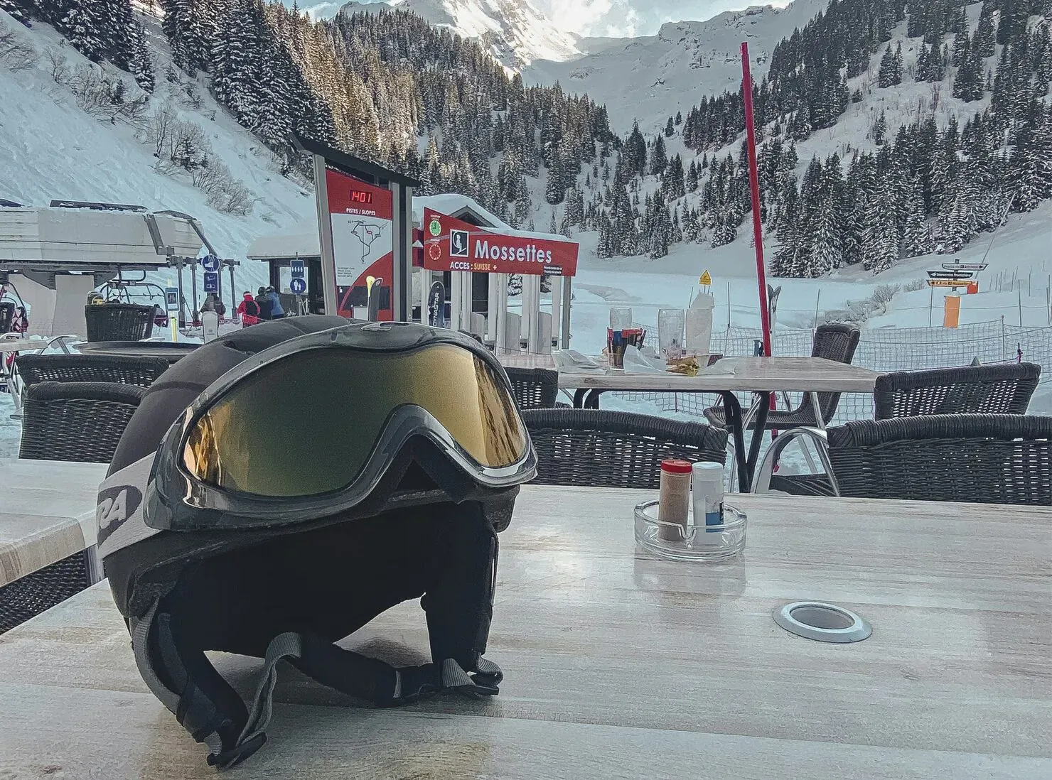 Black ski helmet with gold-tinted goggles on snowy mountain cafe table at Montagneuse resort, alpine backdrop.