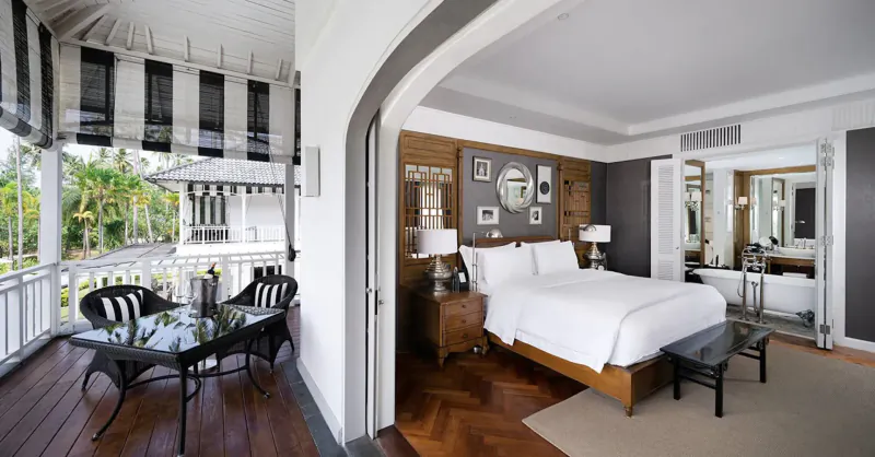 Elegant luxury hotel suite with striped balcony overlooking tropical palms, wicker furniture, king bed, mirrors, and bathtub.