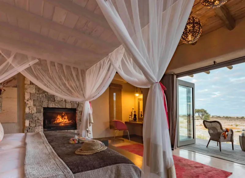 Luxurious king bed with white canopy in Onguma lodge room, stone fireplace lit, open doors to Etosha savanna view.