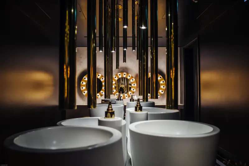 Luxurious gallery bathroom with white sinks, gold hanging pipes, and glowing circular wall lights