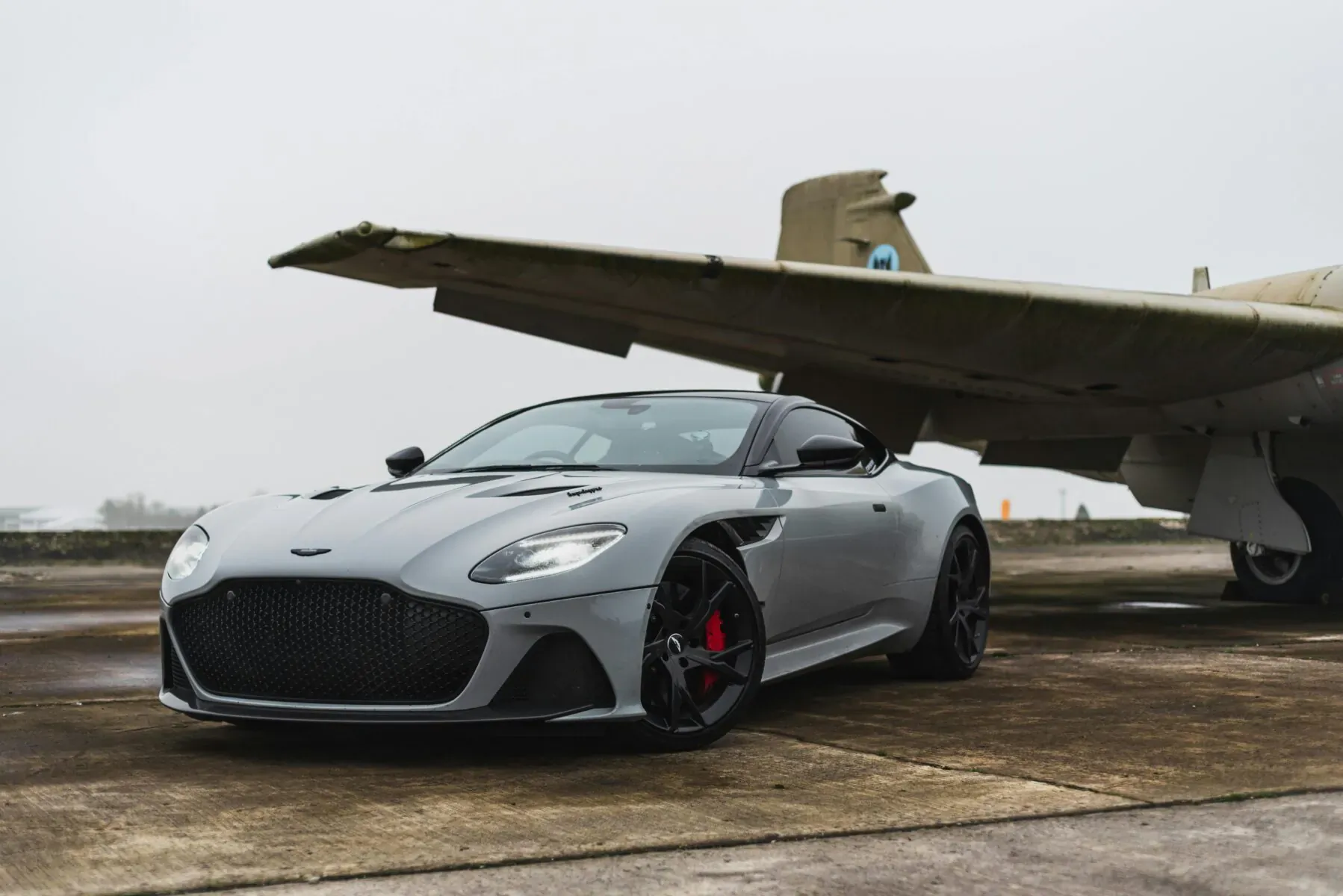 Silver Aston Martin sports car parked under the wing of a vintage airplane on a foggy tarmac.