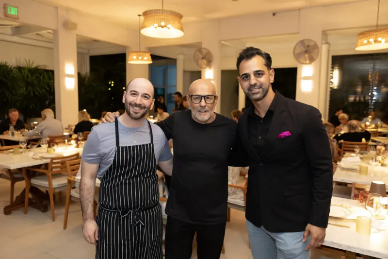 Three men smiling arm-in-arm in upscale restaurant: chef in striped apron, man in black, man in pink-pocketed black blazer.