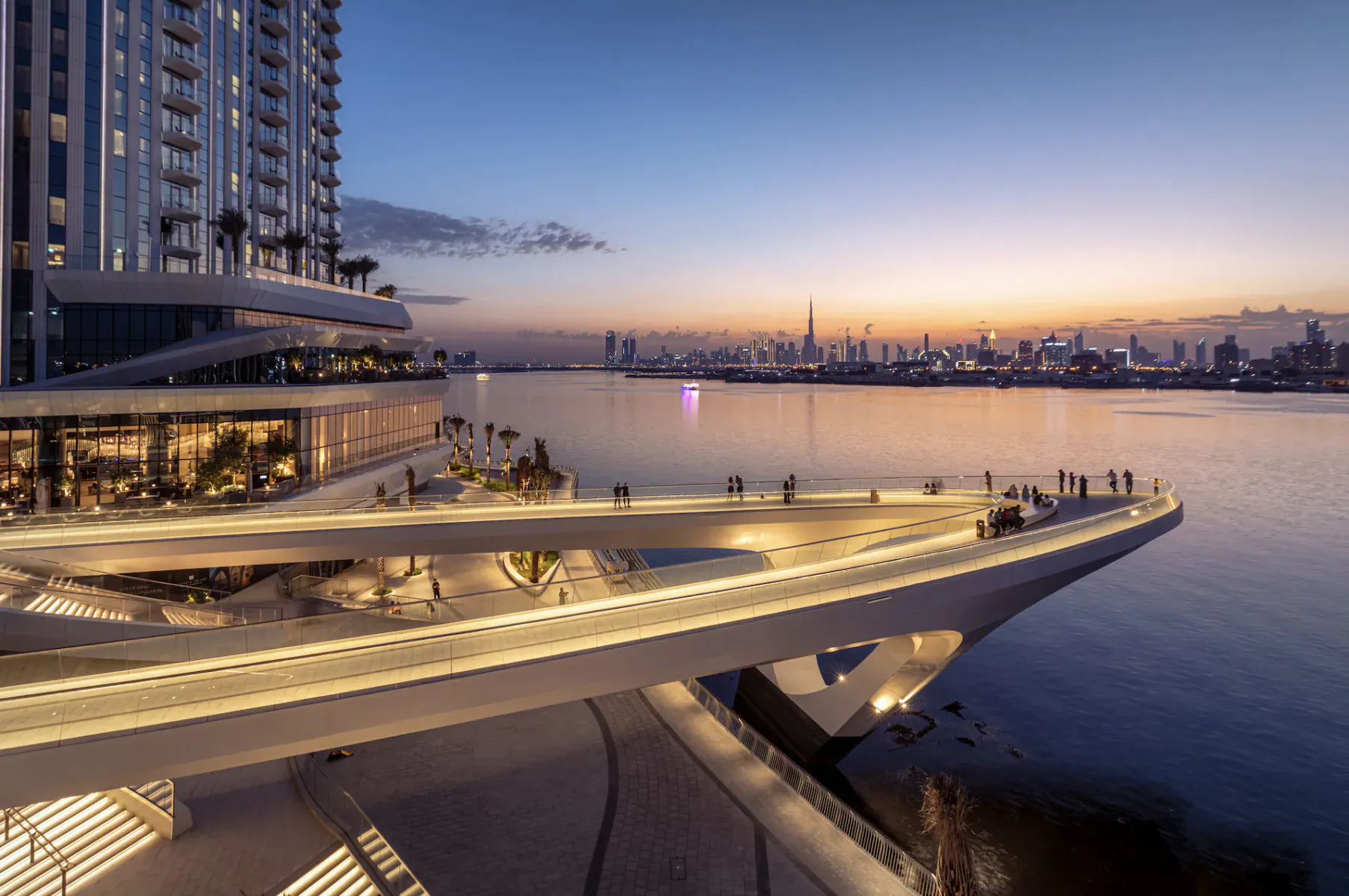 Evening view of Address Grand Creek Harbour: modern high-rise, curved lit skybridge over Dubai Creek with city skyline at sunset.