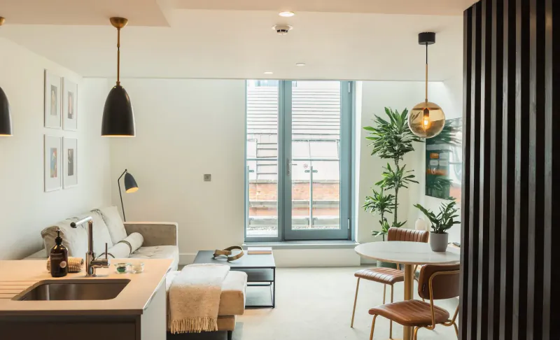 Modern Leven Manchester apartment interior with white sofa, kitchen counter, round dining table, plants, and large window.