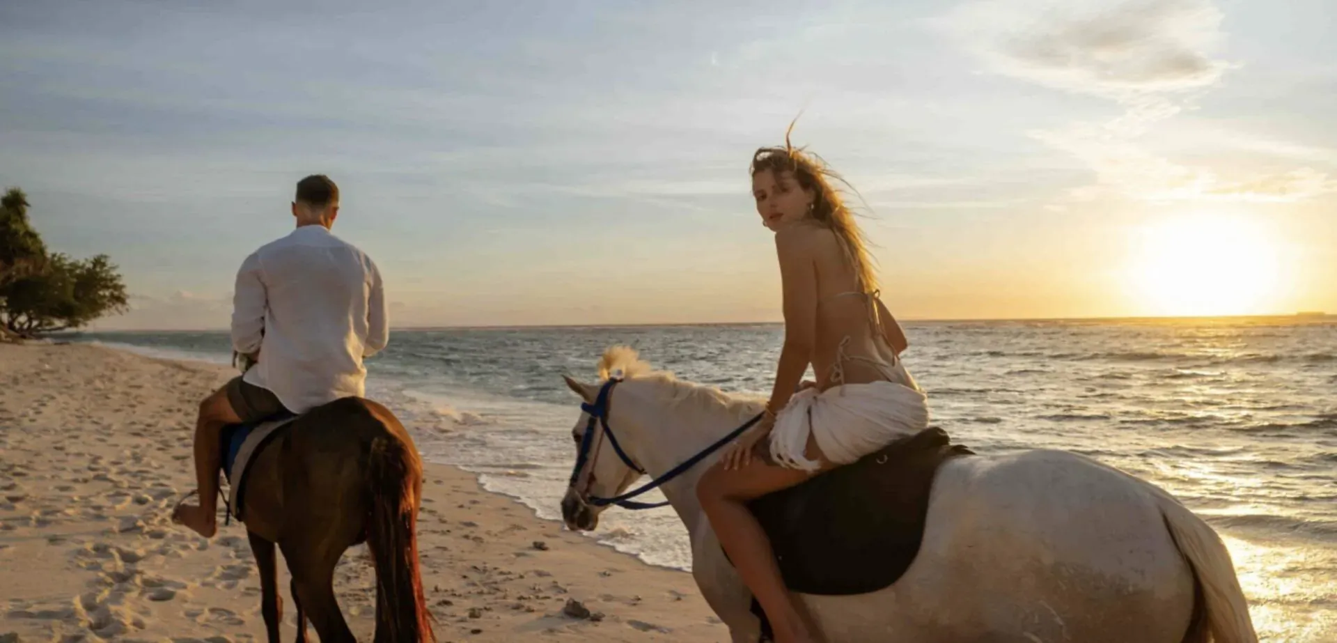 Couple riding horses on Gili Trawangan beach at sunset, man on brown horse, woman on white horse by turquoise sea.