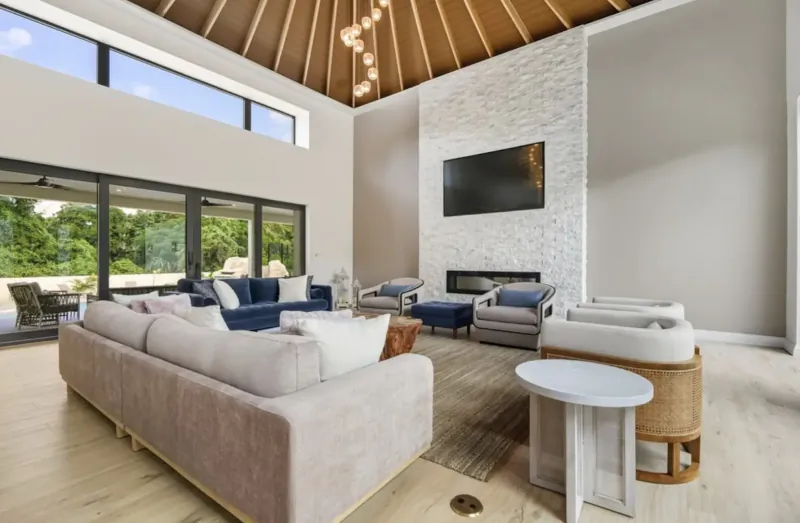 Spacious modern living room in Lyford Cay luxury home with high wood-beamed ceiling, stone fireplace, plush sofas, and garden view.