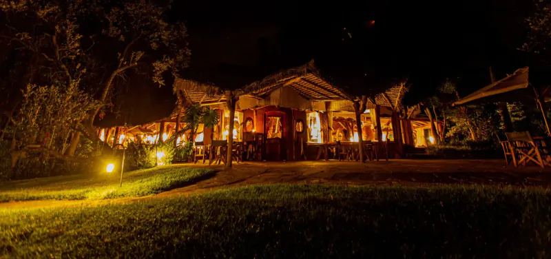 Chui Lodge at night in Lake Naivasha, Kenya: warmly lit thatched building with tables, surrounded by trees and grass.