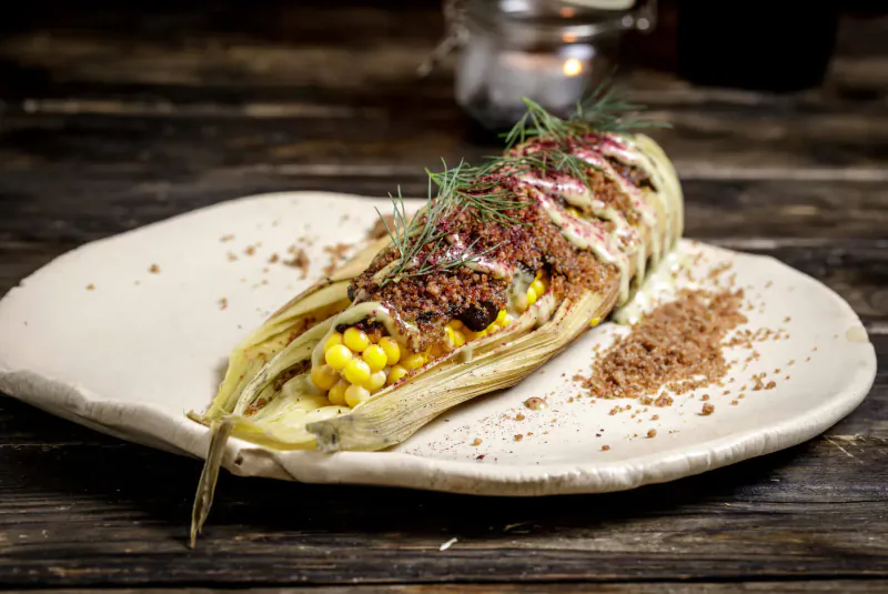 Roasted corn on the cob topped with creamy sauce, red relish, dill, and spice dust on a white plate