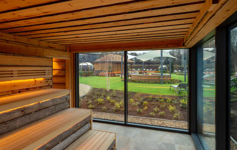 Wooden sauna interior with tiered benches, large windows overlooking lush spa garden at Carden Park.