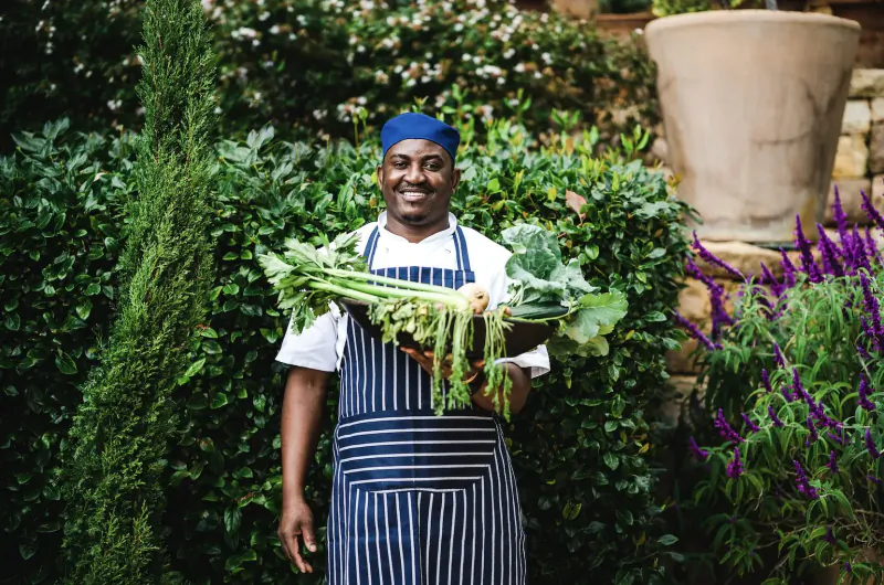 Smiling Black chef in blue toque and striped apron holds fresh greens and kale in lush garden at 21 Nettleton.