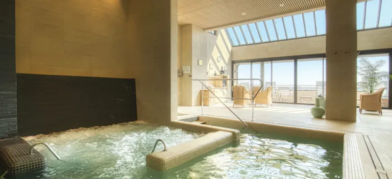 Luxurious indoor hot tub with bubbling water in modern spa, wooden walls, lounge chairs, and panoramic window view.