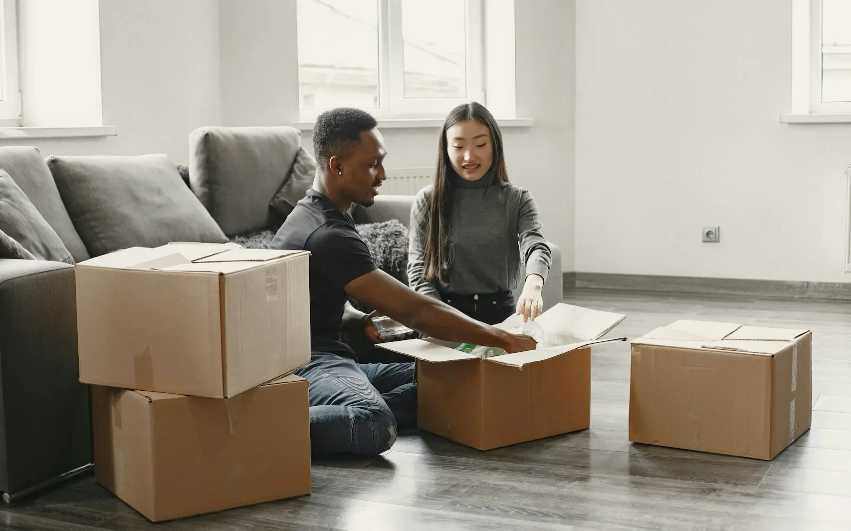 Black man and Asian woman sitting on floor unpacking boxes in modern living room with large windows.