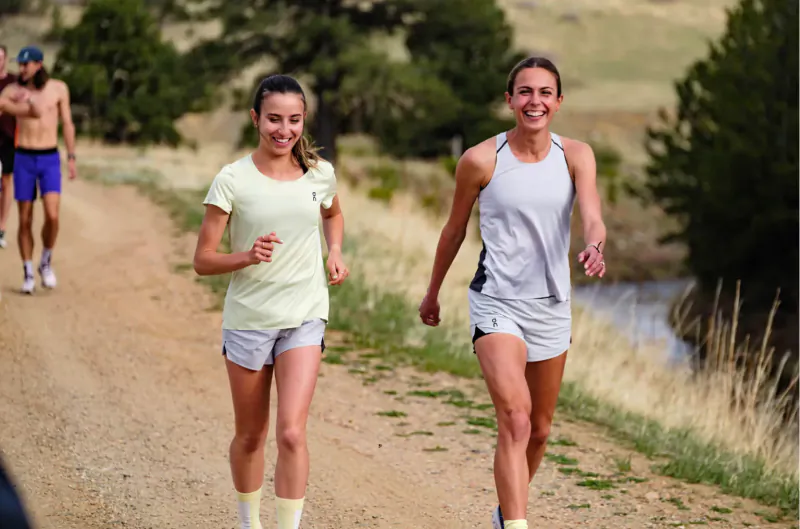 Two smiling women in tank tops and shorts jogging on a dirt trail by a river, with pine trees and hills.