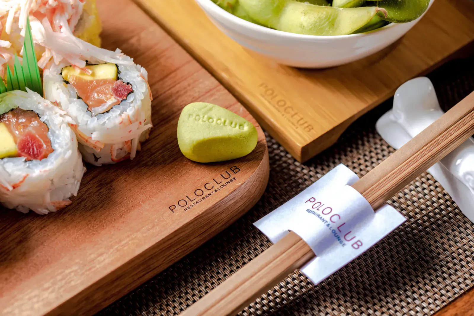 Assorted sushi rolls with avocado and wasabi on Polo Club wooden board, chopsticks, and edamame bowl