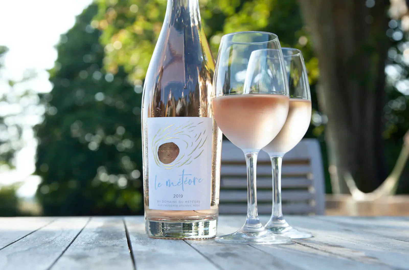 Le Météore Rosé wine bottle and two glasses on wooden table outdoors amid greenery