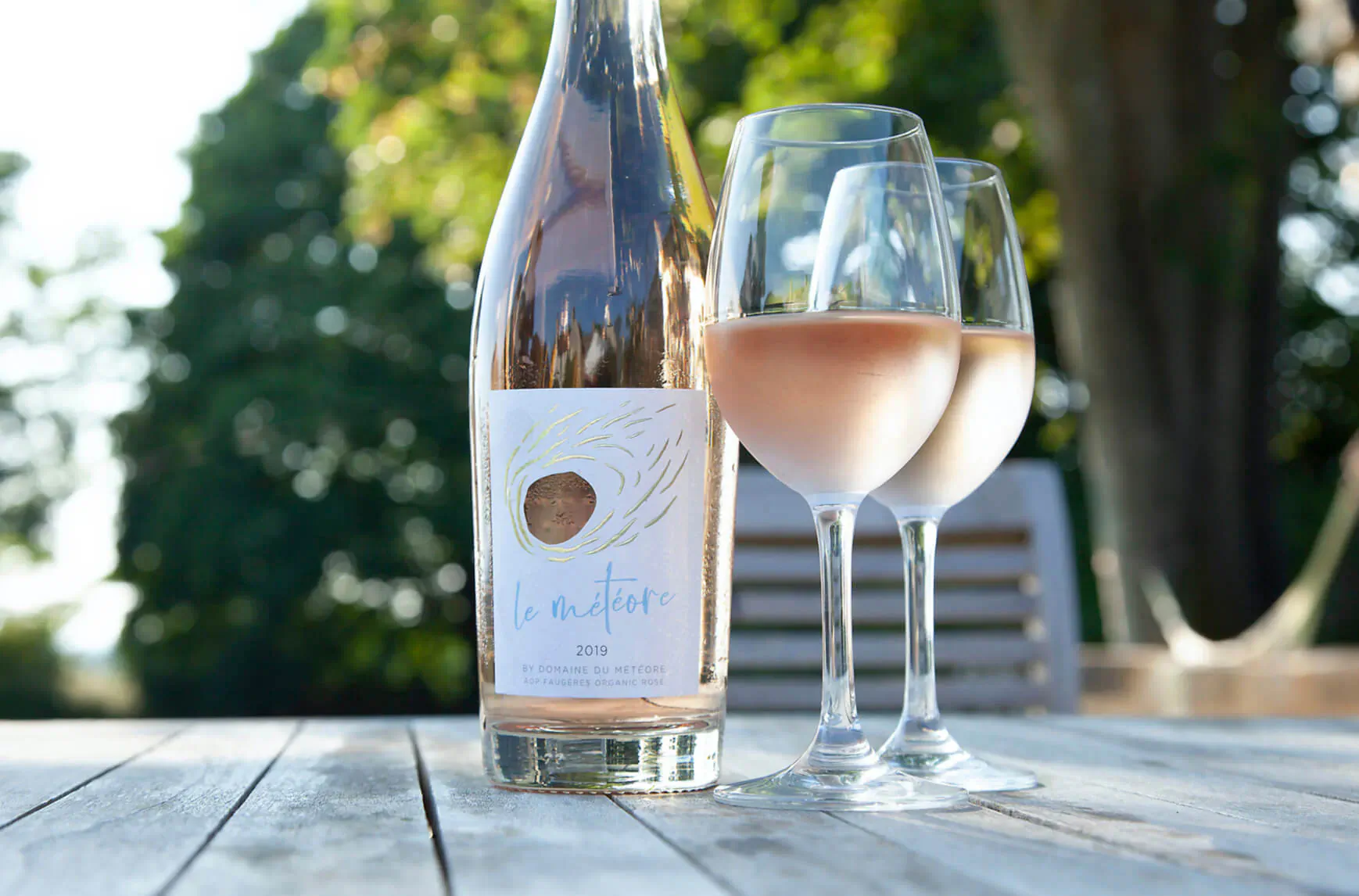 Le Météore Rosé wine bottle and two glasses on wooden table outdoors amid greenery