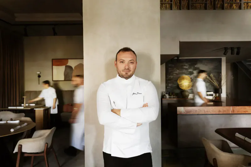 Chef in white uniform arms crossed, standing confidently in modern Makris Restaurant kitchen with staff blurred in background.