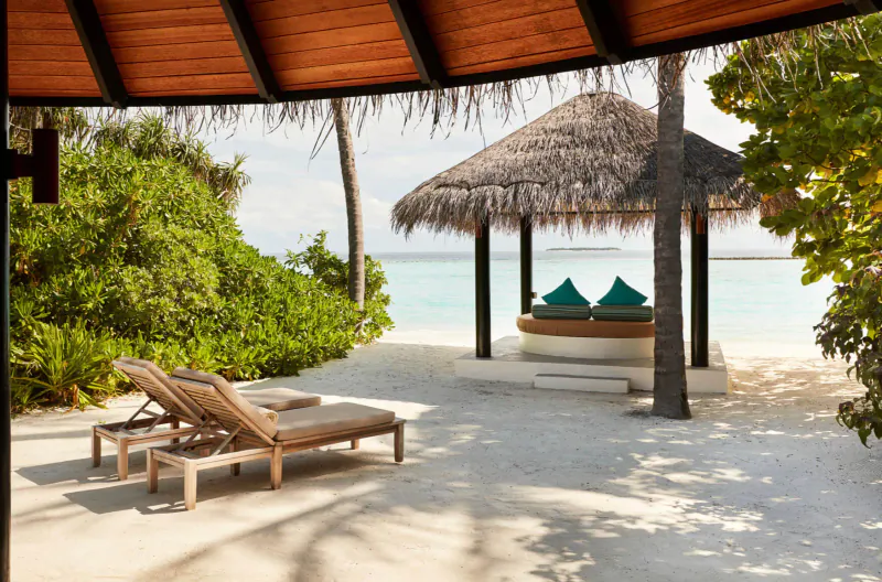 Open beachside pavilion with thatched roof, turquoise cushions on circular bench, two wooden loungers on white sand, tropical greenery and ocean.