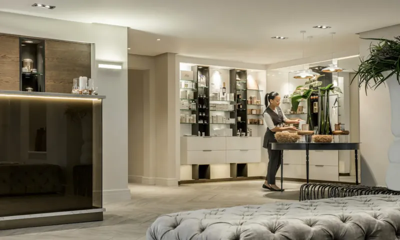 Hotel receptionist in uniform stands behind modern reception desk with glassware, plants, and ottoman in elegant lobby.