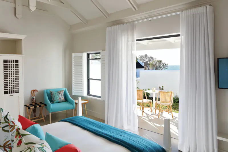 Bright bedroom at The Old Rectory with teal bed, pillows, armchair, and open doors to ocean-view patio.