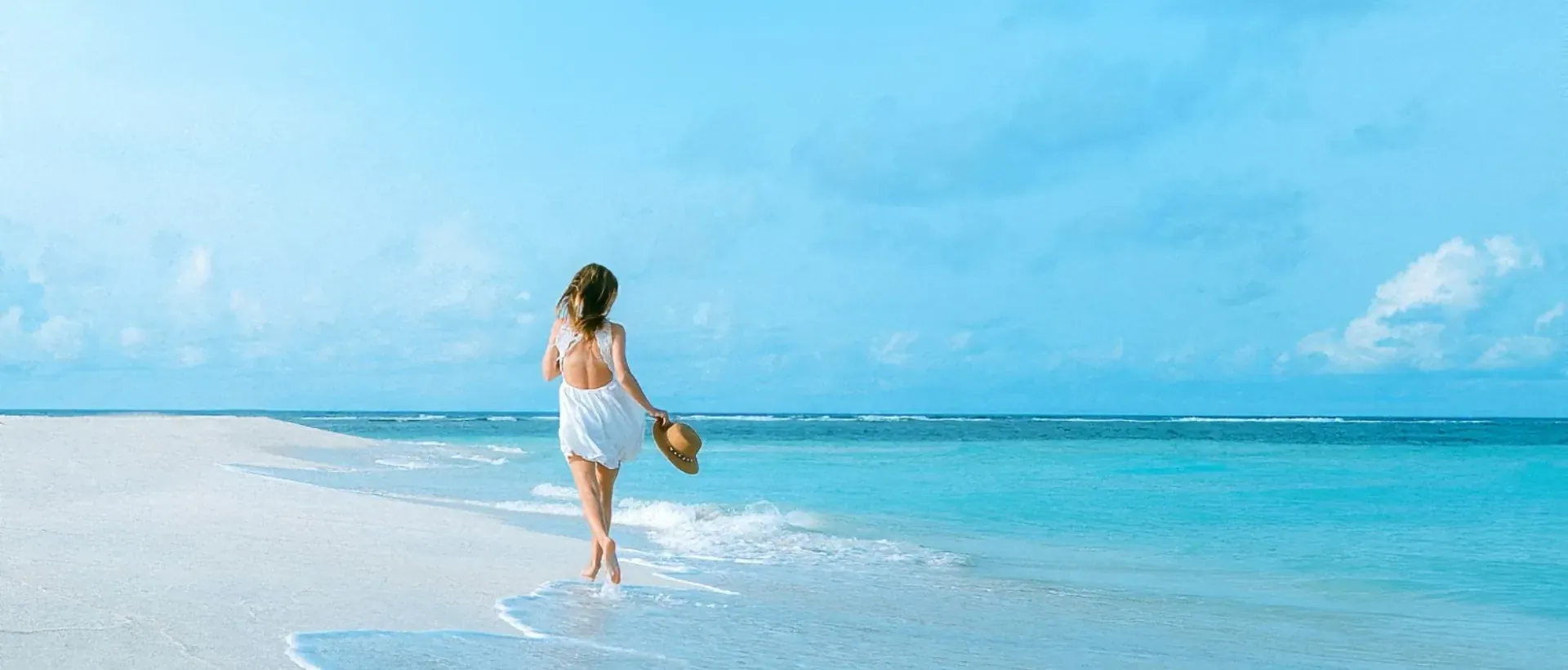 Woman in white dress and hat walks barefoot on white sandy beach toward turquoise ocean under blue sky
