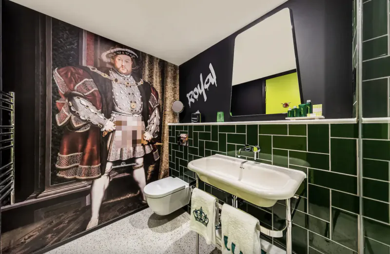 Stylish hotel bathroom with large mural of Henry VIII in Tudor attire on wall, green tiles, white sink, toilet, and towels.