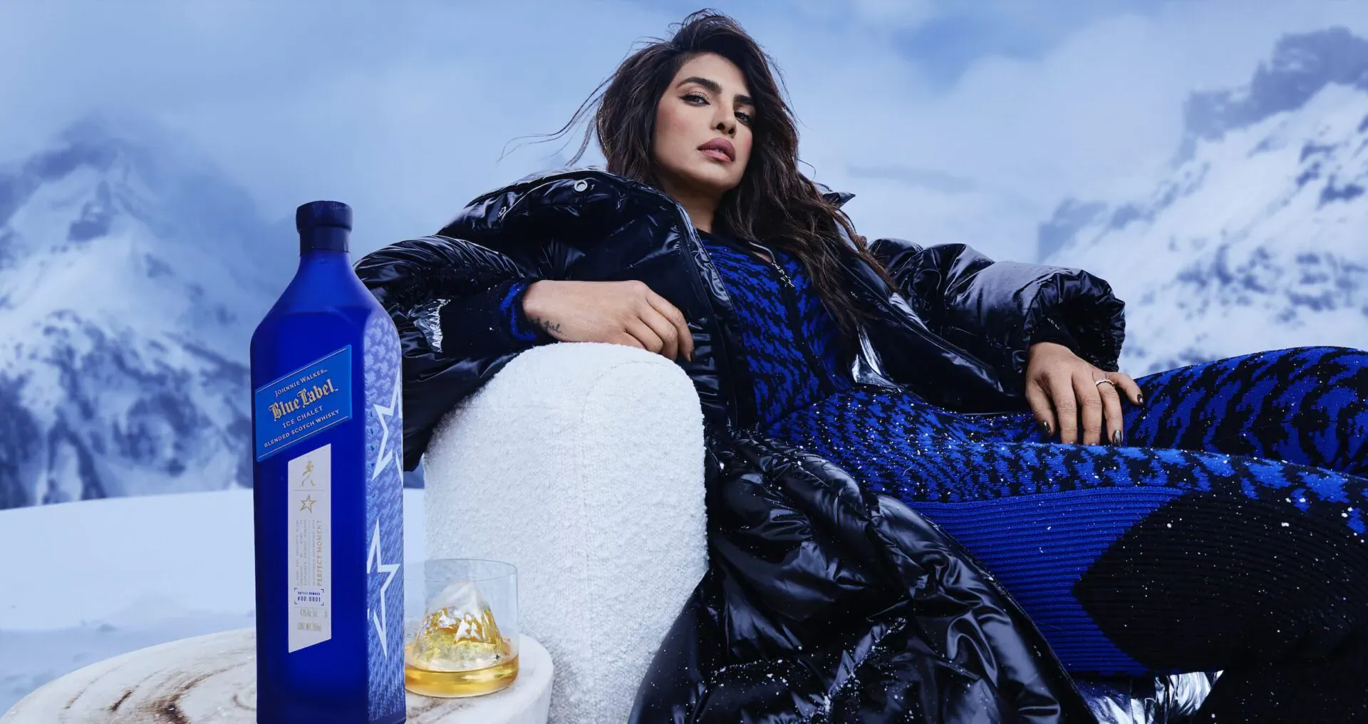Priyanka Chopra lounging in fur coat on snowy mountain with Johnnie Walker Blue Label bottle and glass beside white chair, après ski.