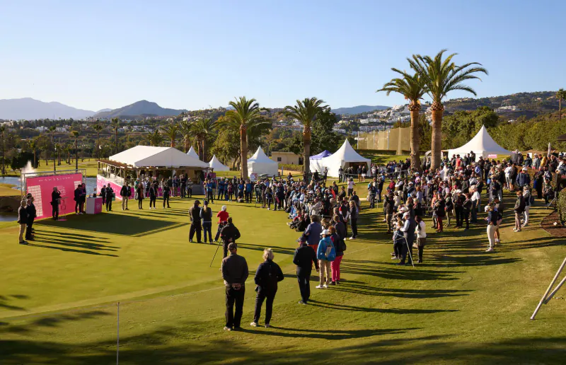 Crowd gathers near pink and white tents on green golf course at Los Naranjos, Marbella, with palm trees and mountains.