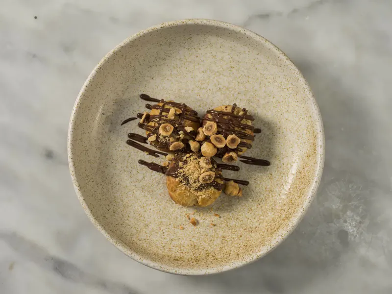 Three hazelnut pastries topped with chocolate drizzle on a textured white plate