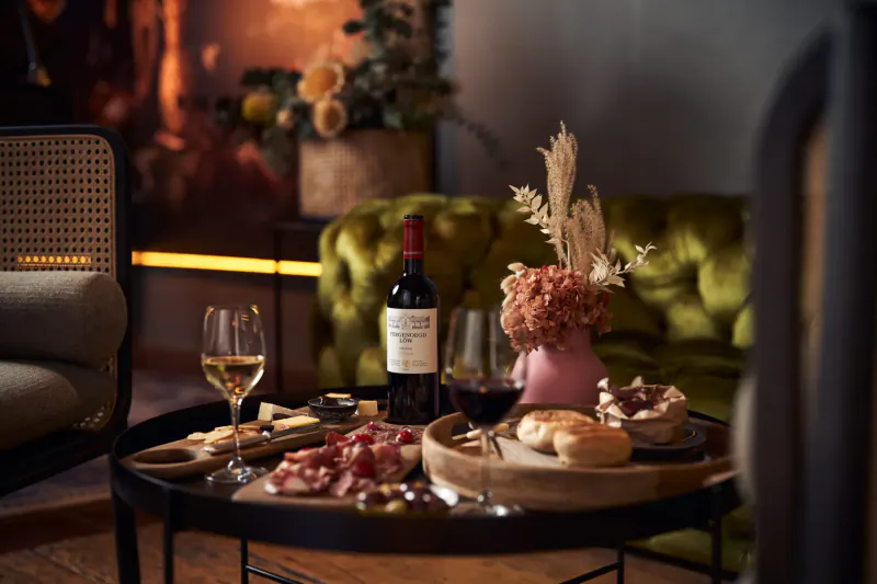 Elegant charcuterie board with cheeses, meats, breads on wooden tray, red wine bottle, white wine glasses, pink flowers on green velvet sofa in stylish setting