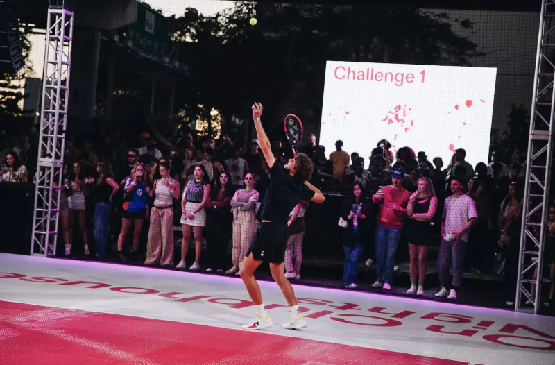 Male athlete in black shorts raises racket triumphantly on red court during Challenge 1 event, cheering crowd and stage backdrop.