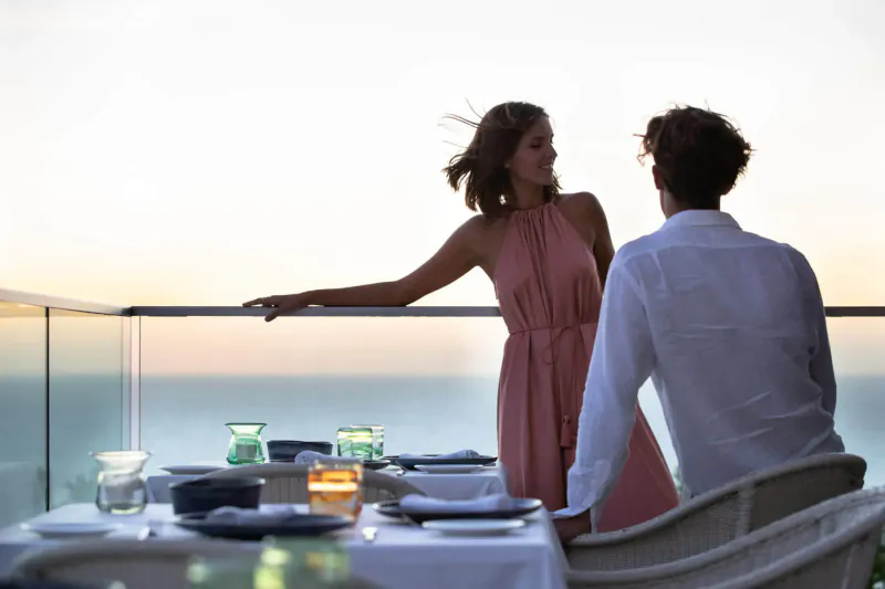 Couple in conversation at candlelit table on balcony overlooking sunset sea at Jumeirah Port Soller Hotel.